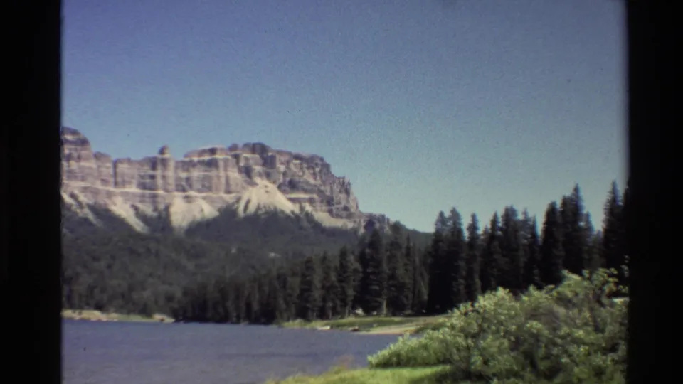 1980 home-movie frame of a Western U.S. mountain range, lake, and pine trees seen through dark 8mm frame edges.
