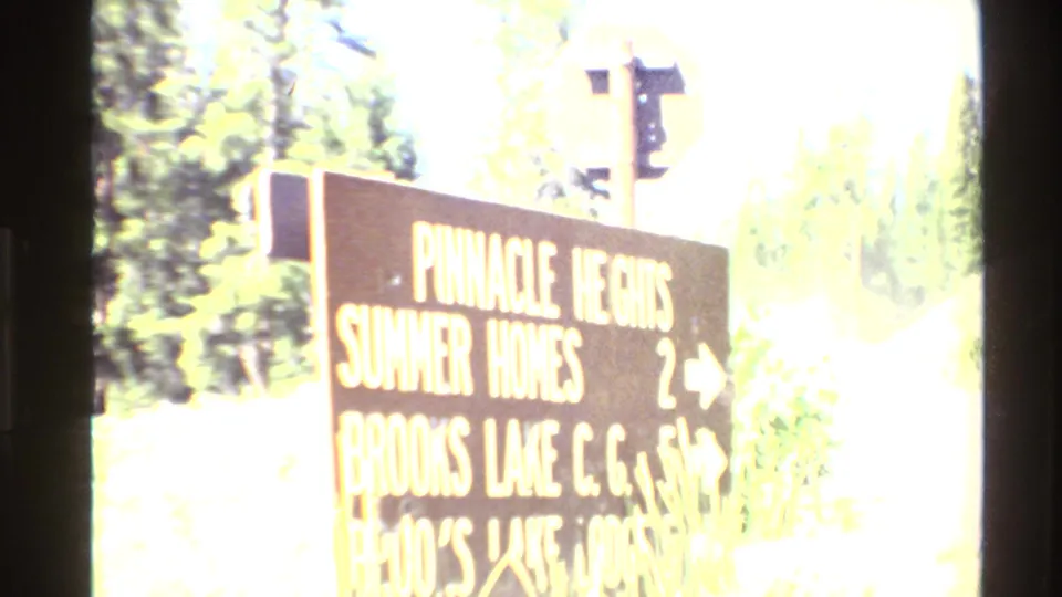 Wooden roadside sign pointing to Pinnacle Heights, Summer Homes, and Brooks Lake C.G. in a bright mountain setting.