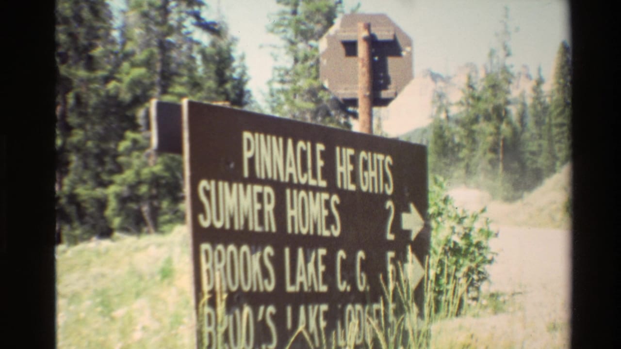 1980s United States 8mm frame showing a brown sign for Pinnacle Heights Summer Homes and Brooks Lake destinations beside a rural road.