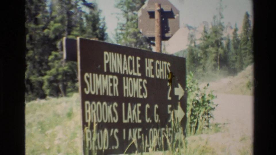 Soft 8mm frame showing the same brown vacation-area directional sign, partly obscured by motion blur and grass in the foreground.