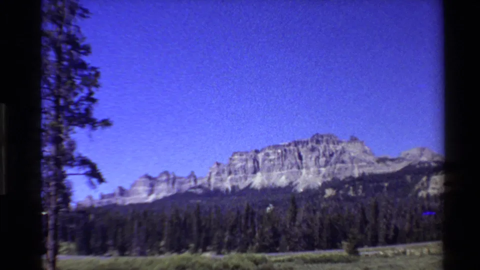 1980 Western USA home-movie frame showing pale rugged peaks, evergreen forest, and a bright saturated blue sky.