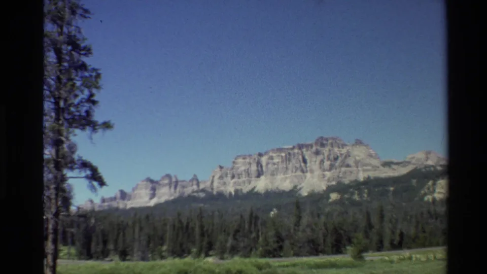 1980 Western U.S. 8mm frame with a broad mountain ridge, dark evergreen forest, open pasture, and clear blue sky.