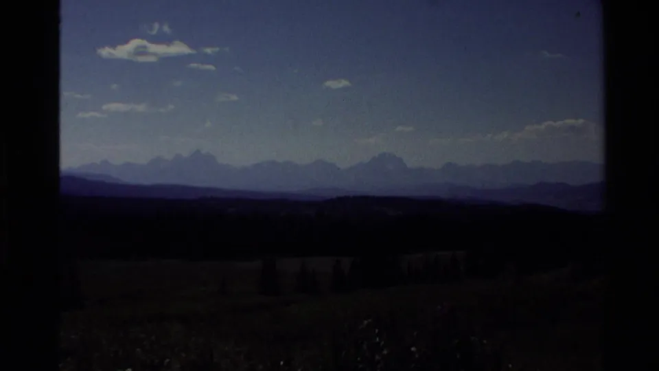 1980 Western U.S. 8mm frame with shadowed foreground land, distant mountain silhouettes, scattered clouds, and deep blue sky.