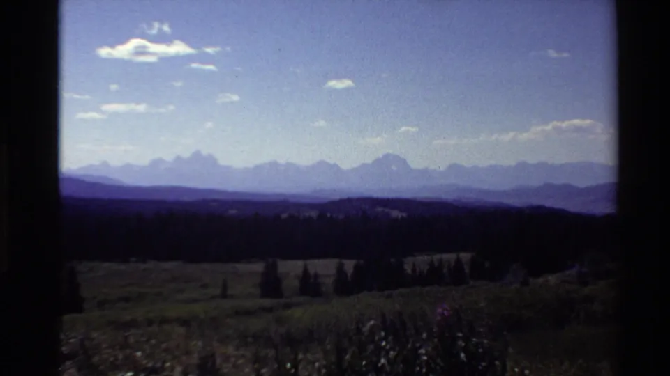 1980 Western U.S. 8mm frame showing distant jagged mountain shapes, dark pine-covered hills, open sky, and a grassy foreground.