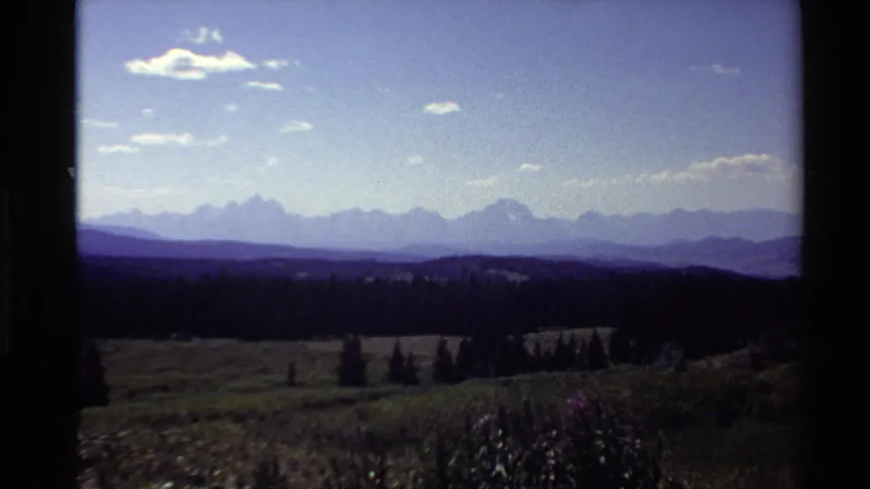 1980 Western U.S. 8mm frame with pale blue sky, scattered clouds, distant mountains, a dark tree line, and low meadow vegetation.