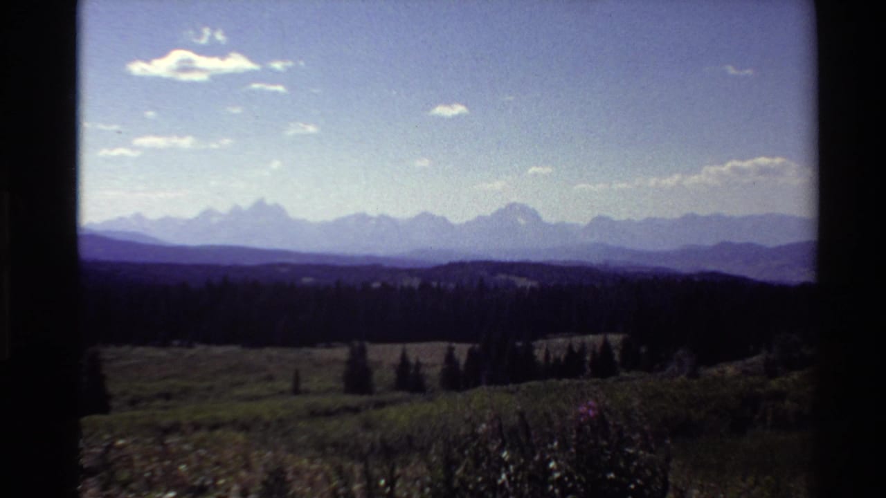 1980 8mm frame showing a distant mountain range, dark evergreen valley, open meadow, and bright sky from an elevated viewpoint.