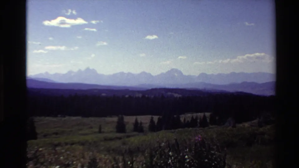 1980 8mm frame showing a distant mountain range, dark evergreen valley, open meadow, and bright sky from an elevated viewpoint.