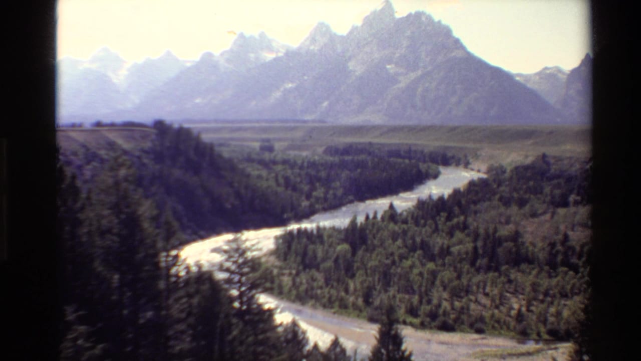 Winding river below forested slopes with jagged mountains in the distance.