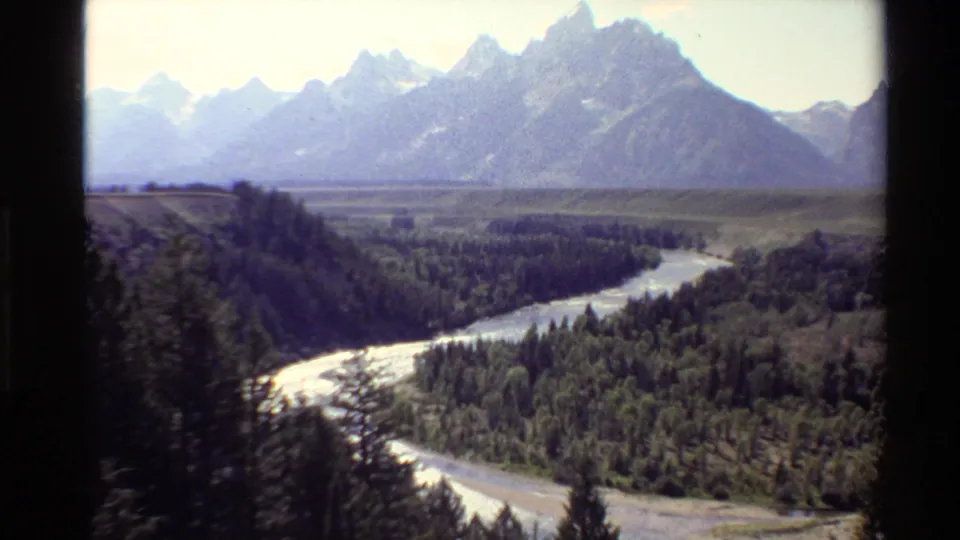 1980 8mm frame showing a pale winding river, dense evergreen trees, open valley, and snow-dusted mountain peaks in the background.