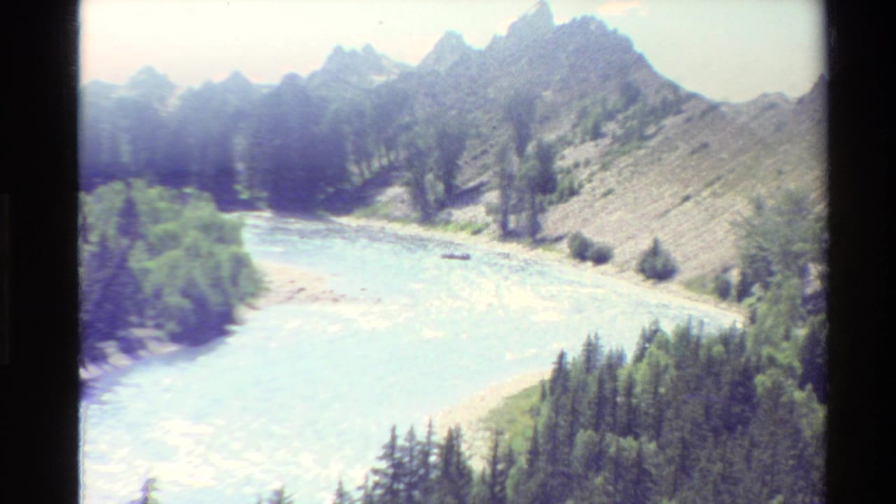 1980s 8mm home-movie frame of a rushing river below evergreen slopes in a rugged Western U.S. mountain valley.