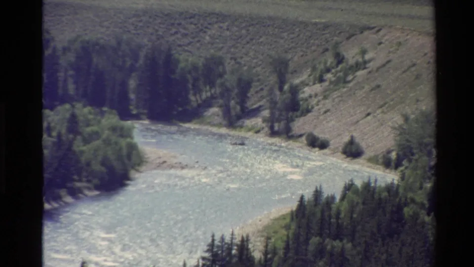 Soft 8mm home-movie view of a turbulent mountain river running between evergreen banks and scrubby slopes.