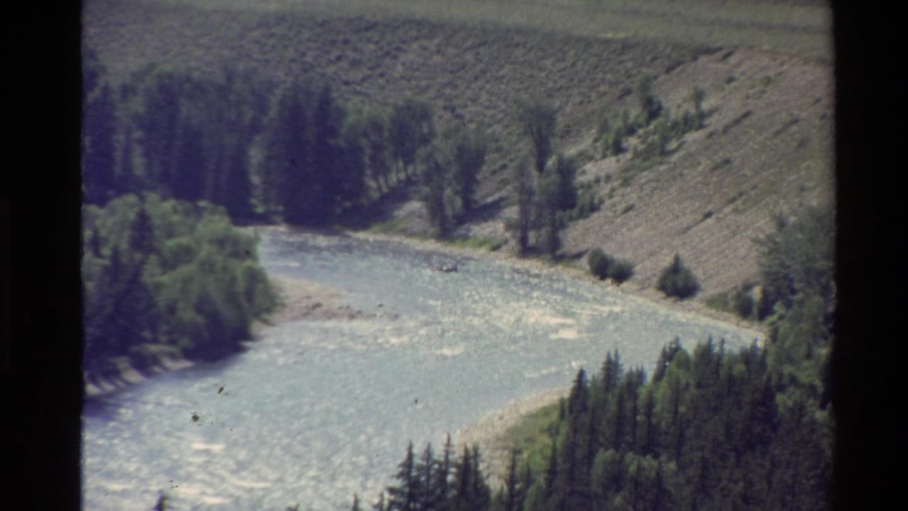 1980s 8mm frame from the Western United States showing a pale river bend, trees, grassy banks, and dry rolling terrain.