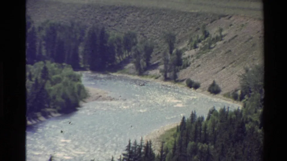 1980s 8mm frame from the Western United States showing a pale river bend, trees, grassy banks, and dry rolling terrain.
