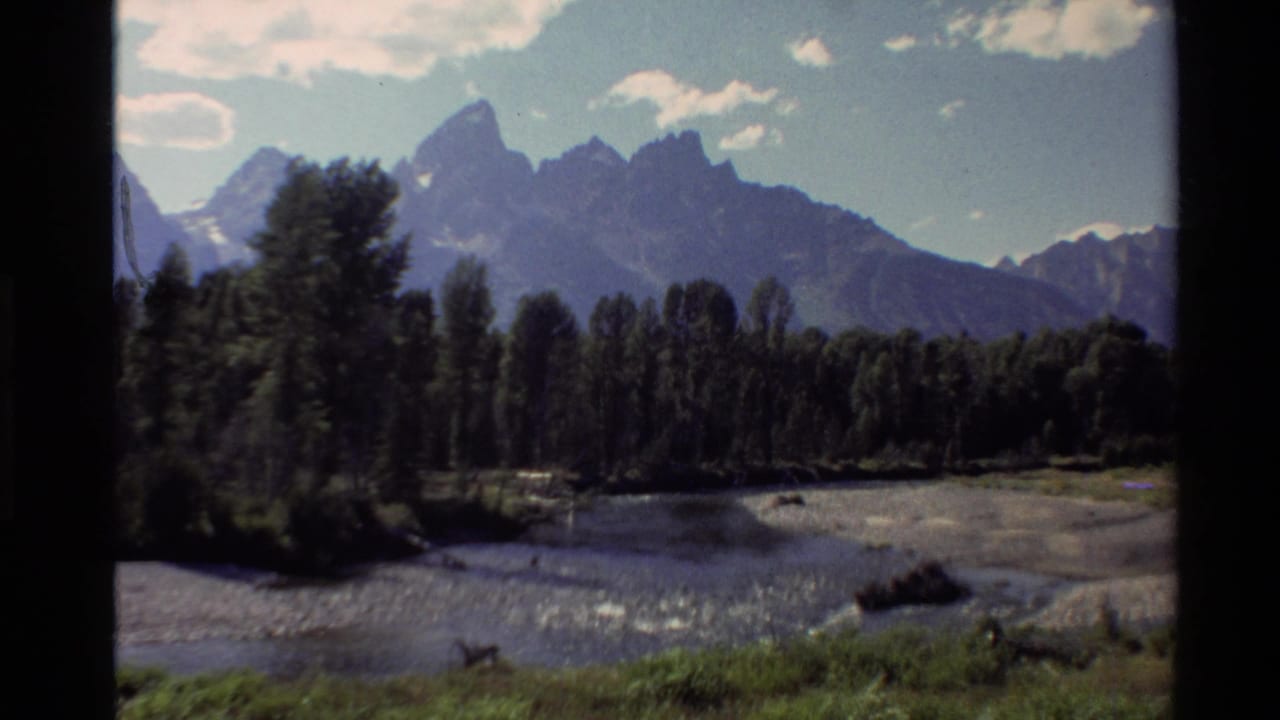 Western U.S. 8mm landscape frame with a jagged mountain range, dark tree line, river channel, and partly cloudy sky.