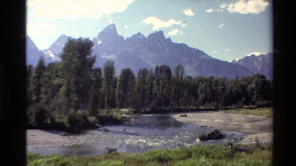 1980s Western U.S. home-movie frame showing a river foreground, dense trees, bright clouds, and distant sharp mountain peaks.