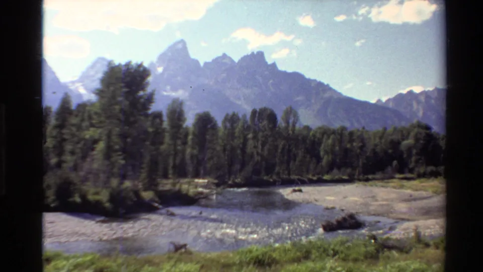 Western U.S. 8mm frame with green riverbank foreground, a shallow river bend, trees, and blue-gray mountains in the distance.