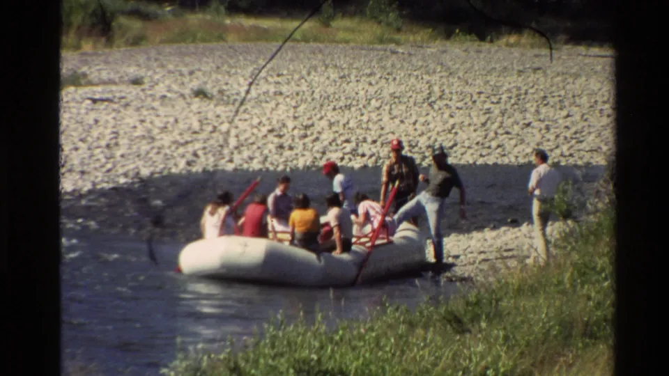 People in casual 1980s clothing gather around a white inflatable raft on a Western U.S. riverbank.