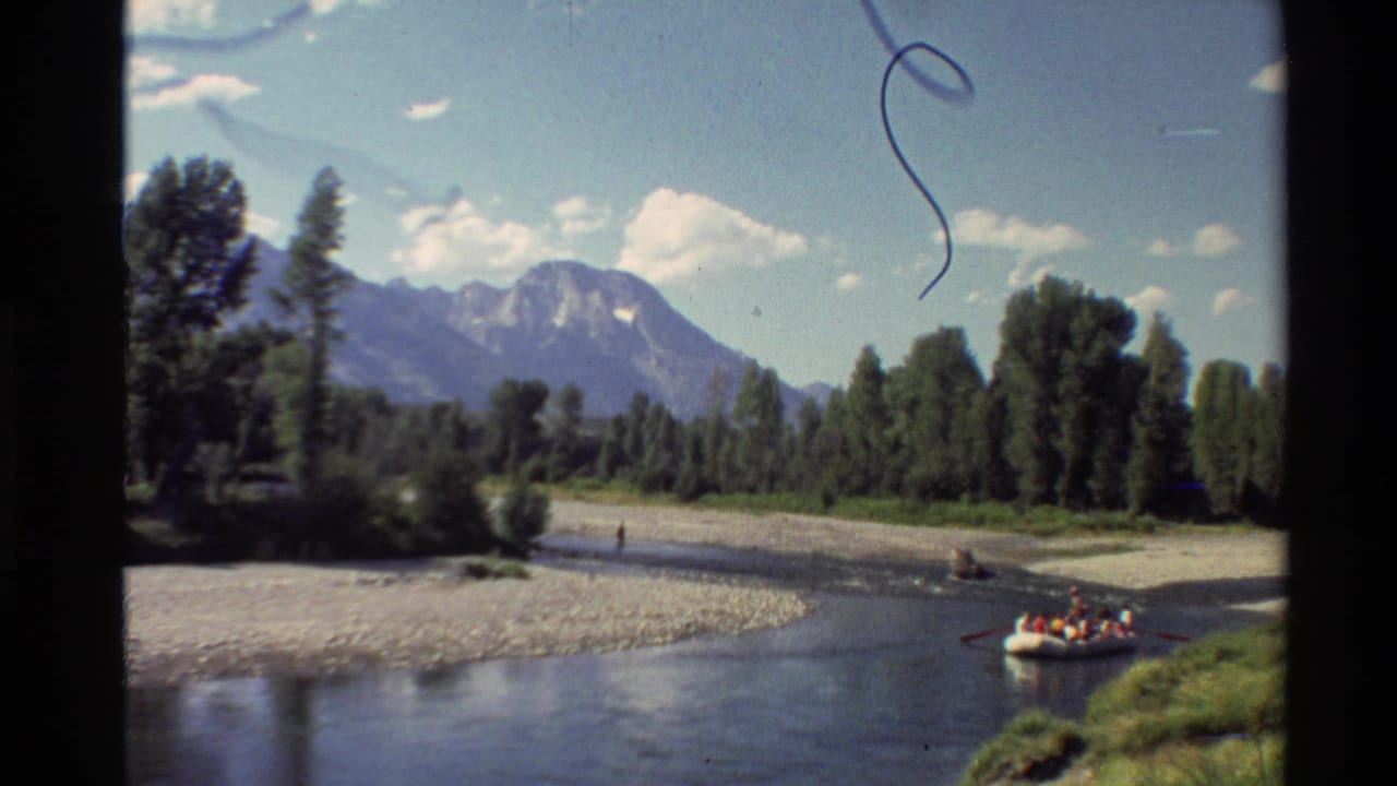 1980 Western USA 8mm view of a calm river below mountains, with trees, gravel banks, and people in a yellow raft.