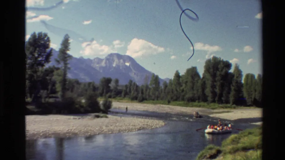 1980 Western USA 8mm view of a calm river below mountains, with trees, gravel banks, and people in a yellow raft.