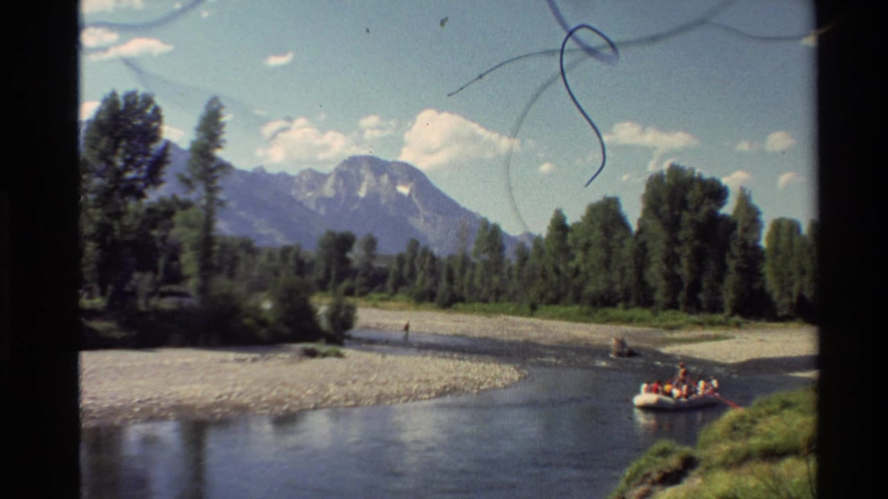 Yellow raft near the right riverbank with mountains, trees, gravel, and sky behind it.