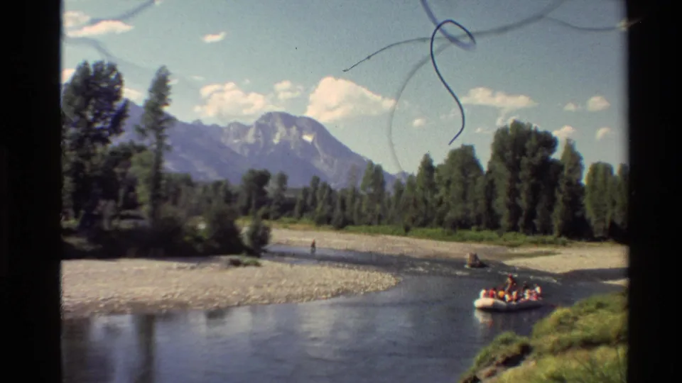 1980 home-movie river view with a walker on the gravel bank and a group in a yellow raft below distant mountains.