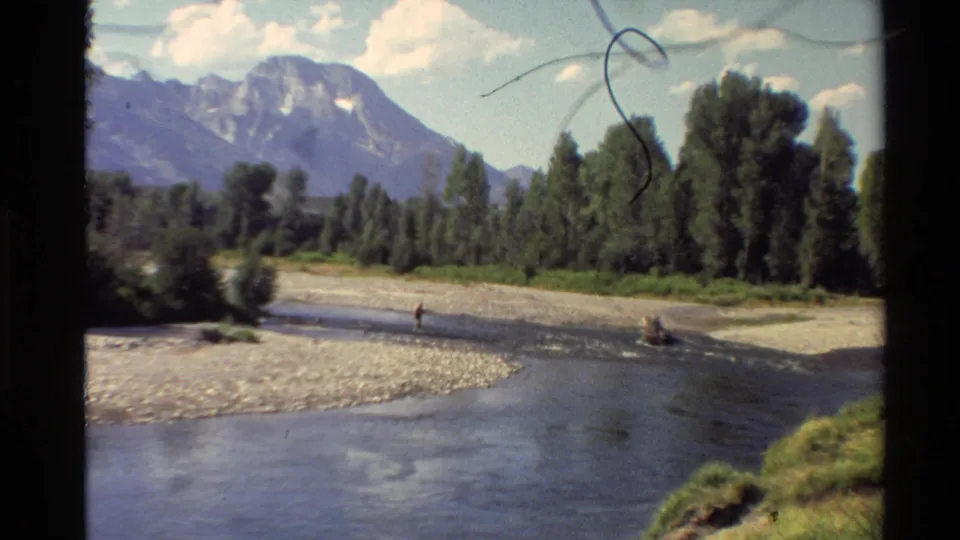 Western USA 8mm mountain river scene showing a distant walker, gravel banks, tree line, and soft home-movie film artifacts.
