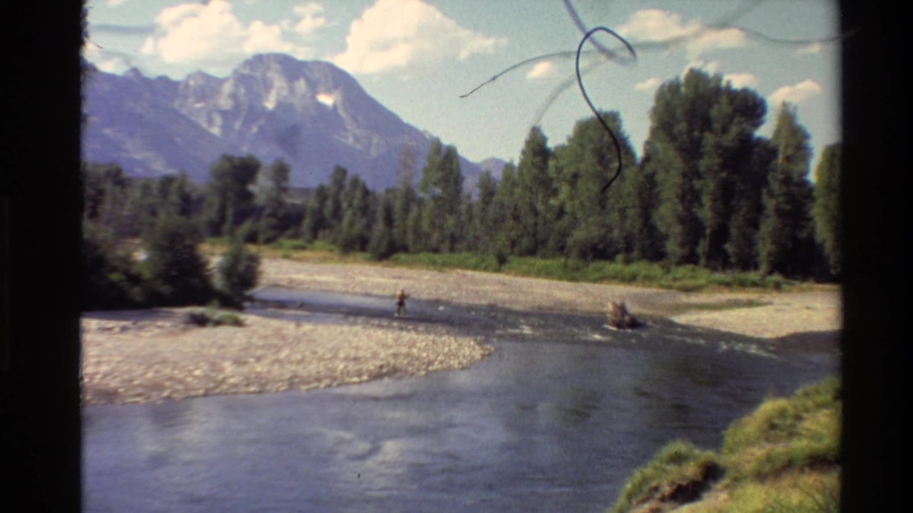 1980 Western U.S. 8mm frame showing a distant person fly fishing in a shallow river below trees and mountains.