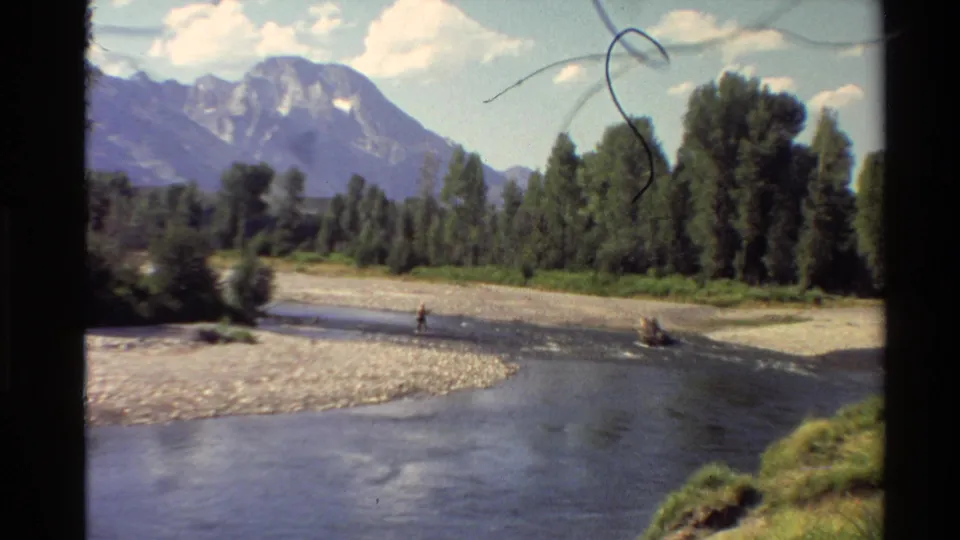 1980 Western U.S. 8mm frame showing a distant person fly fishing in a shallow river below trees and mountains.