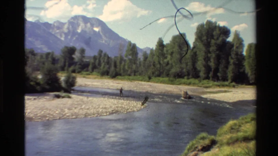 1980 Western U.S. home-movie frame of a clear mountain river with a small fisher and tree-lined bank.