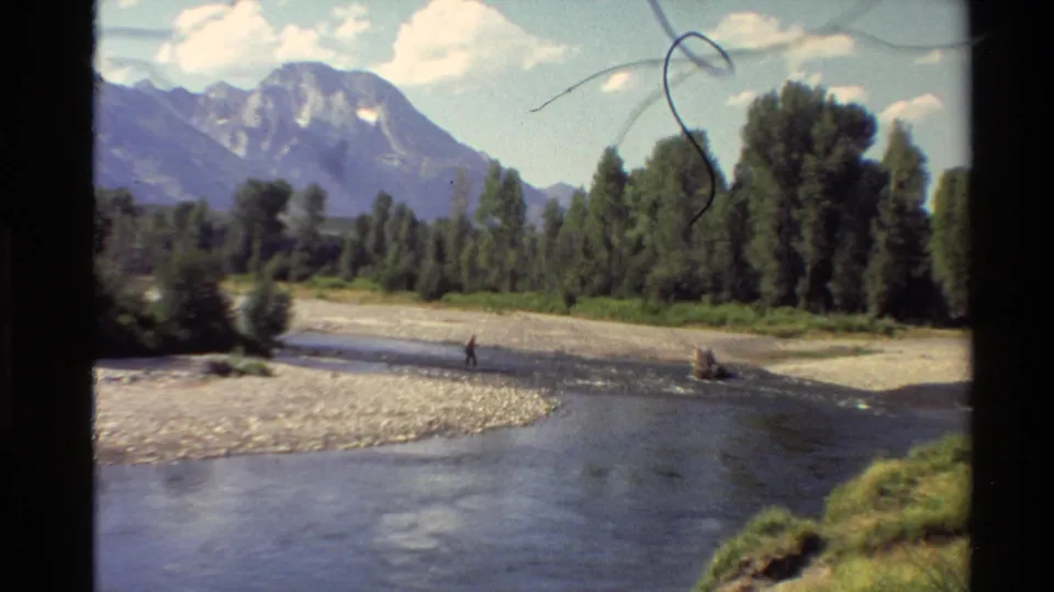 1980 Western U.S. 8mm view of fly fishing on a shallow river framed by trees, mountains, and film-gate edges.