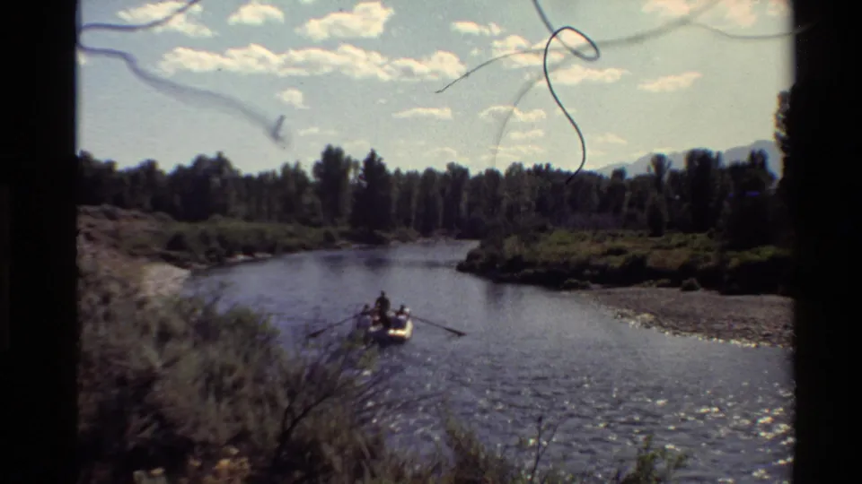 1980 Western U.S. home-movie frame showing people in a small inflatable boat on a clear river below low mountains.