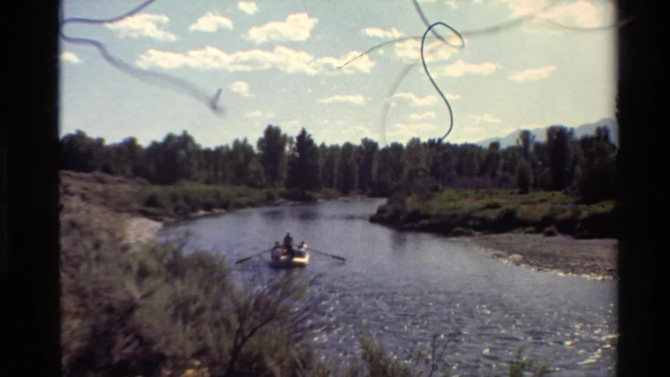 1980 8mm home-movie view of a rowboat on a calm river bend, with wooded banks and a bright clouded sky.