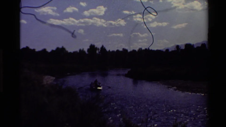 Darkened, purple-tinted 8mm frame showing the rowboat as a silhouette on the river beneath clouds and distant mountains.