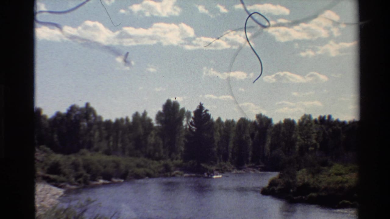 1980 Western U.S. 8mm frame showing a calm river, wooded banks, and scattered clouds, with film dust and gate edges visible.