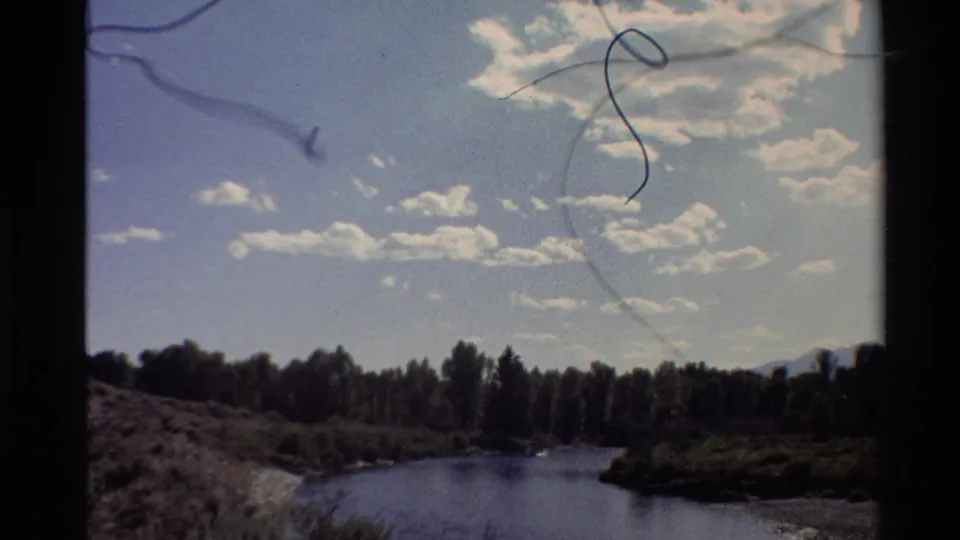1980 Western U.S. 8mm frame of a river bend or inlet bordered by trees under scattered clouds, with dust and scratches in the film image.