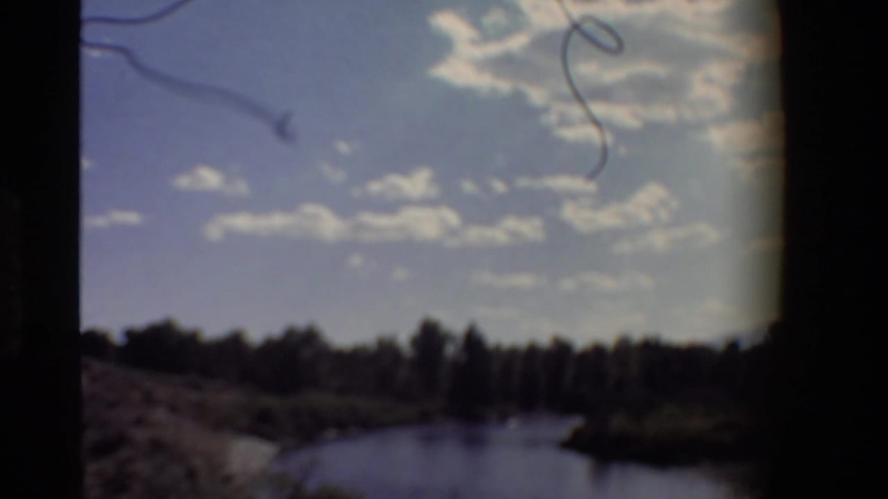 Opening 1980 home-movie frame shows a dim river scene with tree silhouettes, clouds, and visible scratches on the film.