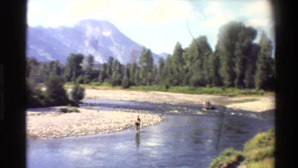 A distant person wades through a shallow Western U.S. river, with a gravel bar, trees, and mountains in the background.
