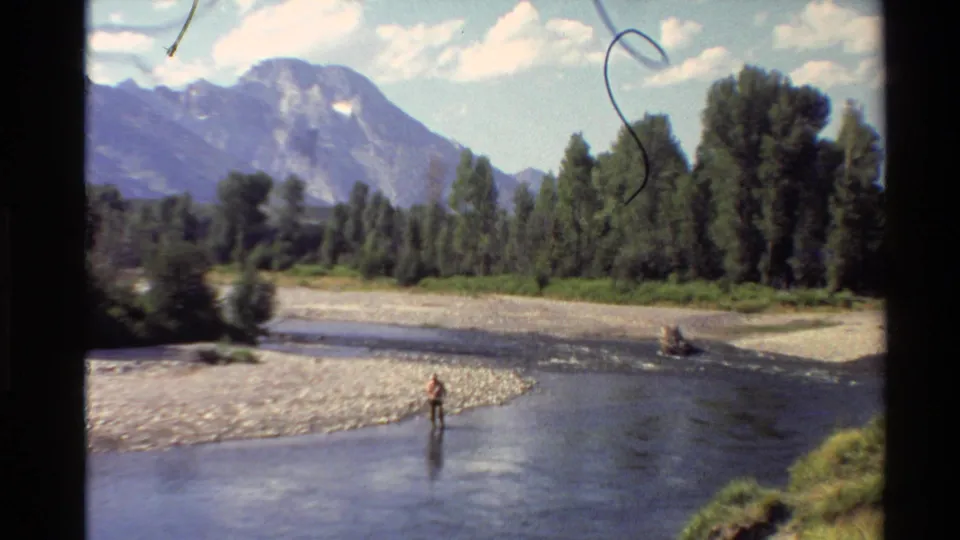 Wide 8mm view of a person in a mountain river, with a rocky bank, tall trees, shrubs, and partly cloudy sky.
