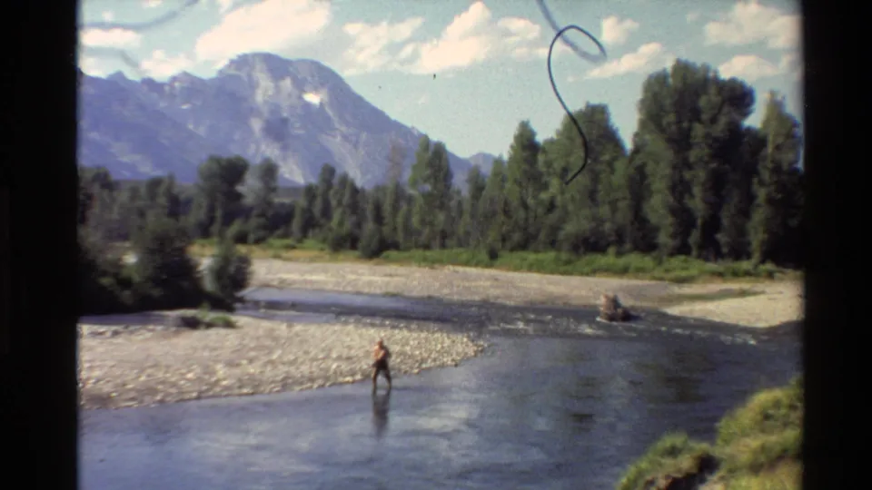 Final frame shows a distant figure in the shallow river, framed by a rocky shore, green trees, mountains, and 8mm film scratches.