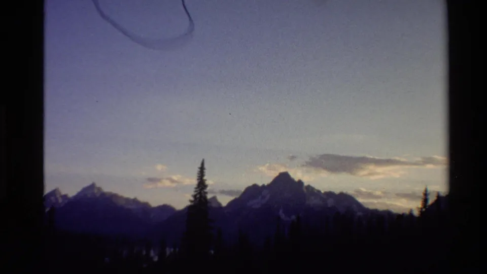 1980 Western U.S. 8mm frame with mountain silhouettes, scattered snow patches, evergreens, and a dimming blue-purple sky.