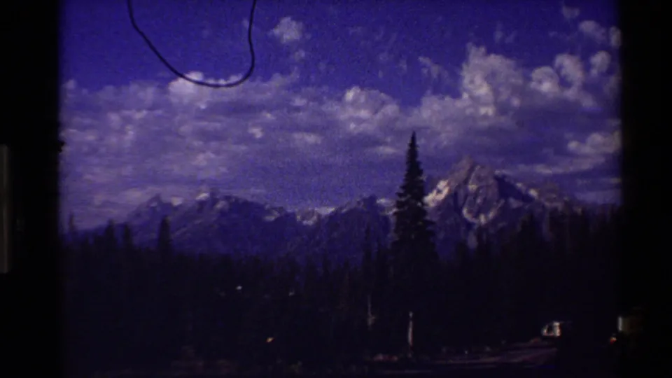 Western U.S. 8mm travel view of distant snow-capped mountains and pine forest beneath a blue sky, partly obscured by dark window framing.