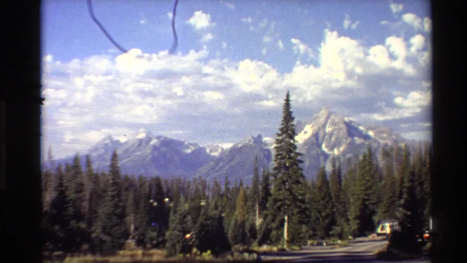 Bright 1980 Western U.S. 8mm frame showing snow-capped peaks, dense pine forest, scattered clouds, and a roadside foreground.