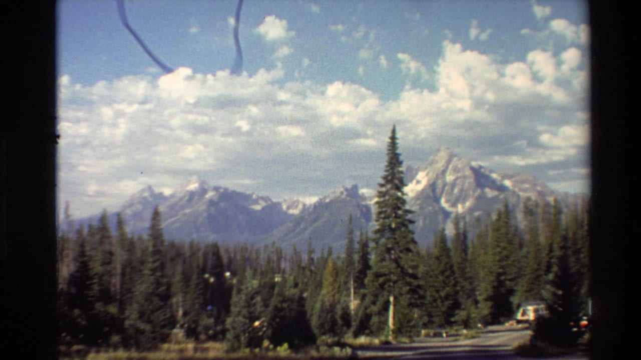 1980s Western USA 8mm frame of evergreen trees before a distant snow-capped mountain range under clouds.