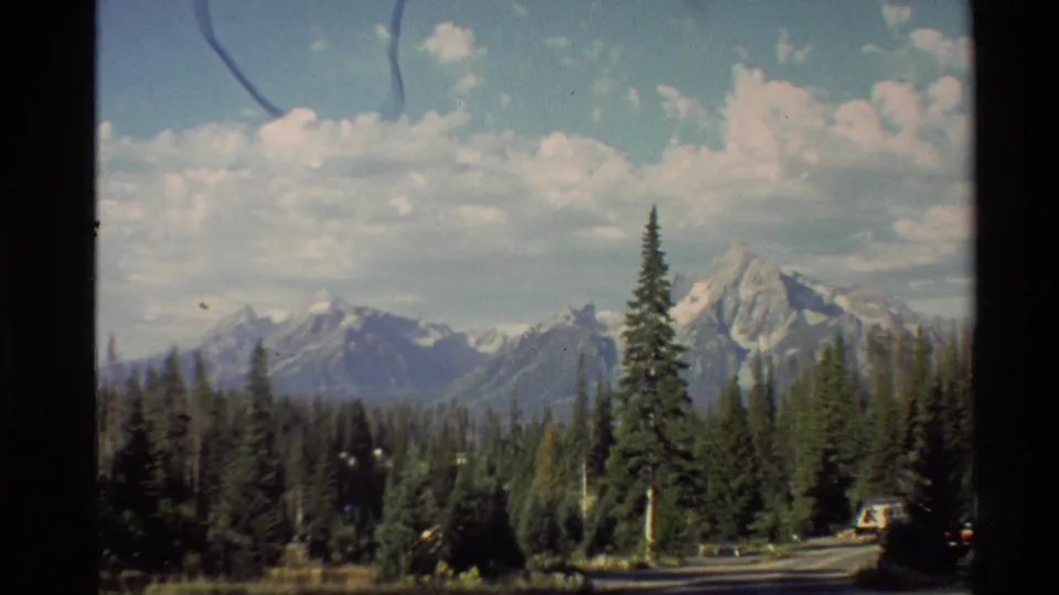 8mm home-movie view of a forested mountain road with snow-capped peaks in the background in the Western USA.