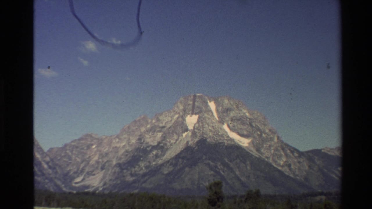 1980 Western U.S. 8mm frame showing rugged mountains with snow patches beneath a clear blue sky and visible film gate borders.