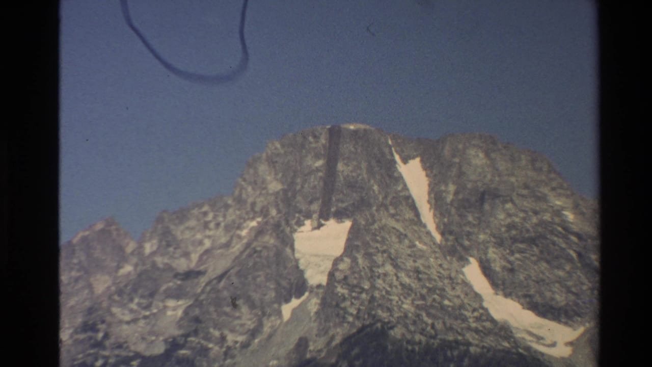 1980 Western USA 8mm frame showing a gray rocky mountain peak, blue sky, snow-filled gullies, and visible film-edge marks.