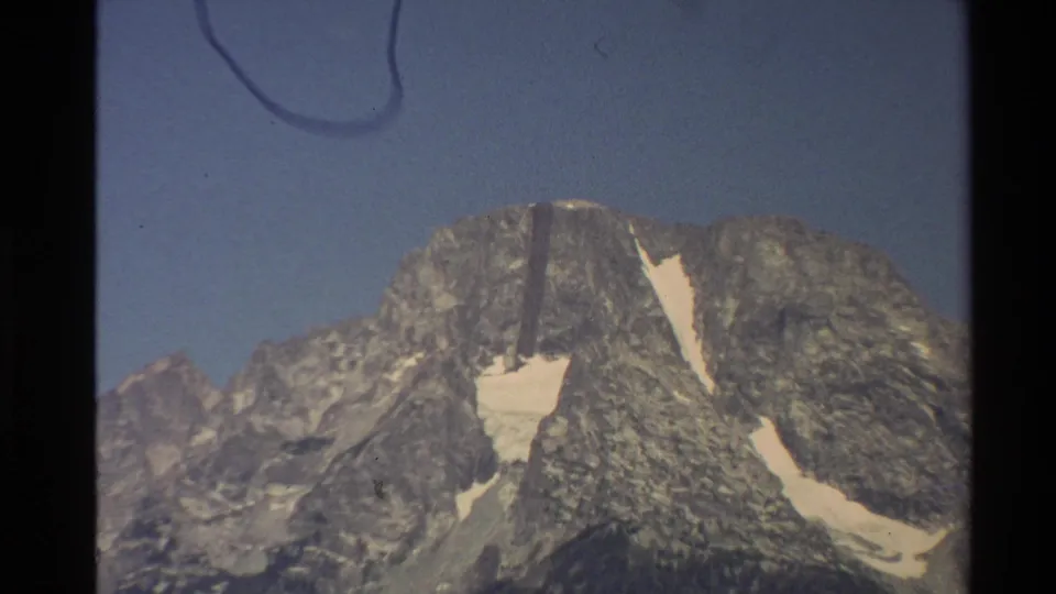 1980 Western USA 8mm frame showing a gray rocky mountain peak, blue sky, snow-filled gullies, and visible film-edge marks.