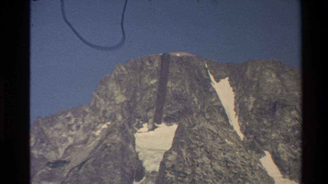 1980 Western USA 8mm view of a rugged gray mountain summit with bright snow patches and a faint curved mark near the top edge.