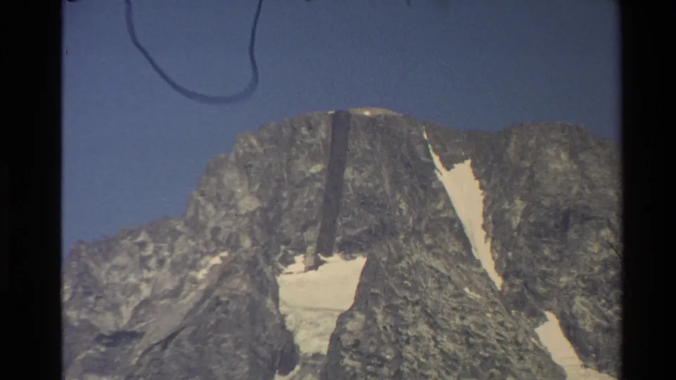 1980 Western USA 8mm view of a rugged gray mountain summit with bright snow patches and a faint curved mark near the top edge.