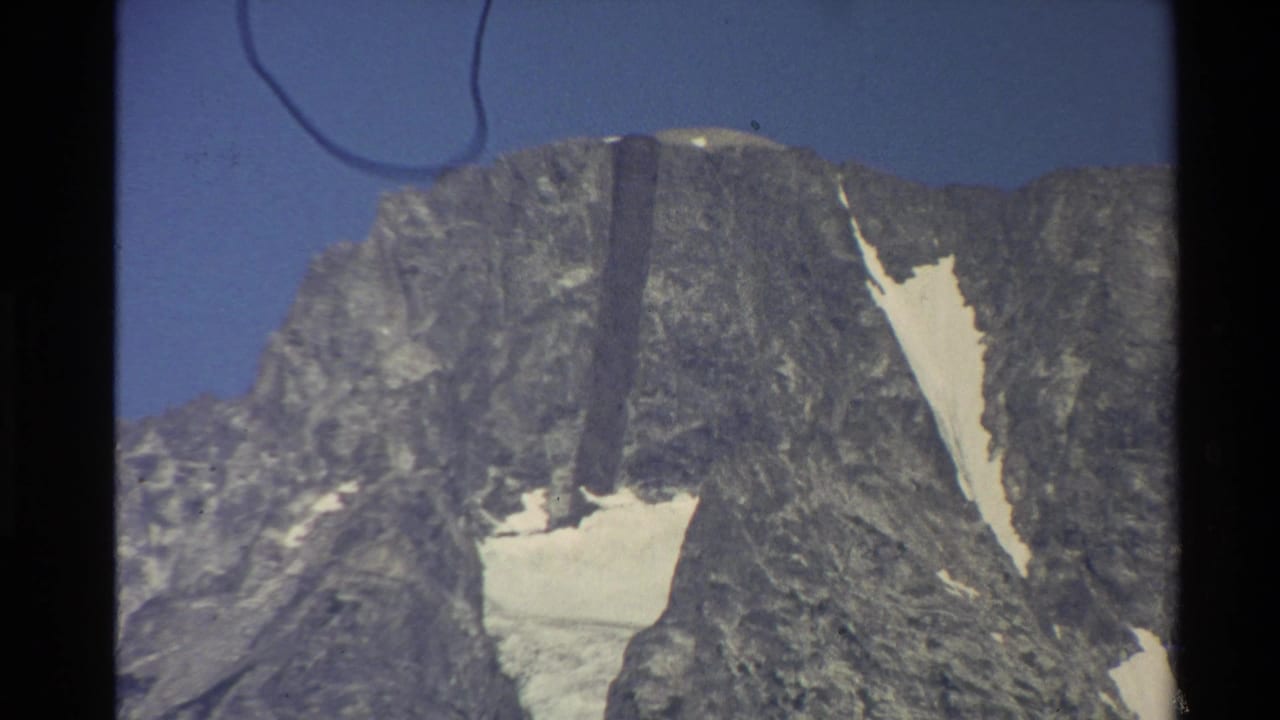 1980 Western U.S. 8mm frame showing a rugged mountain summit with patchy snow and clear sky, with visible film gate edges and dust.